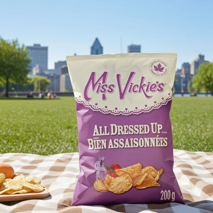 A bag of Miss Vickies All Dressed Up potato chips sits on a checkered picnic blanket in a park, with a blurred city skyline and green lawn in the background.
